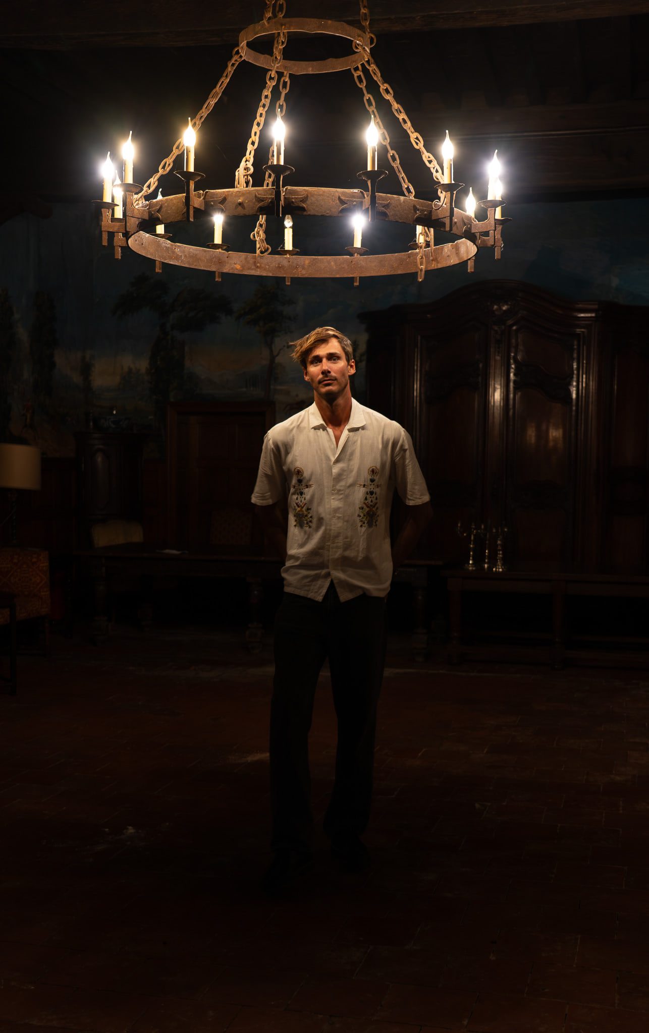 Denver standing beneath a wrought-iron candle chandelier in a panelled château salon.