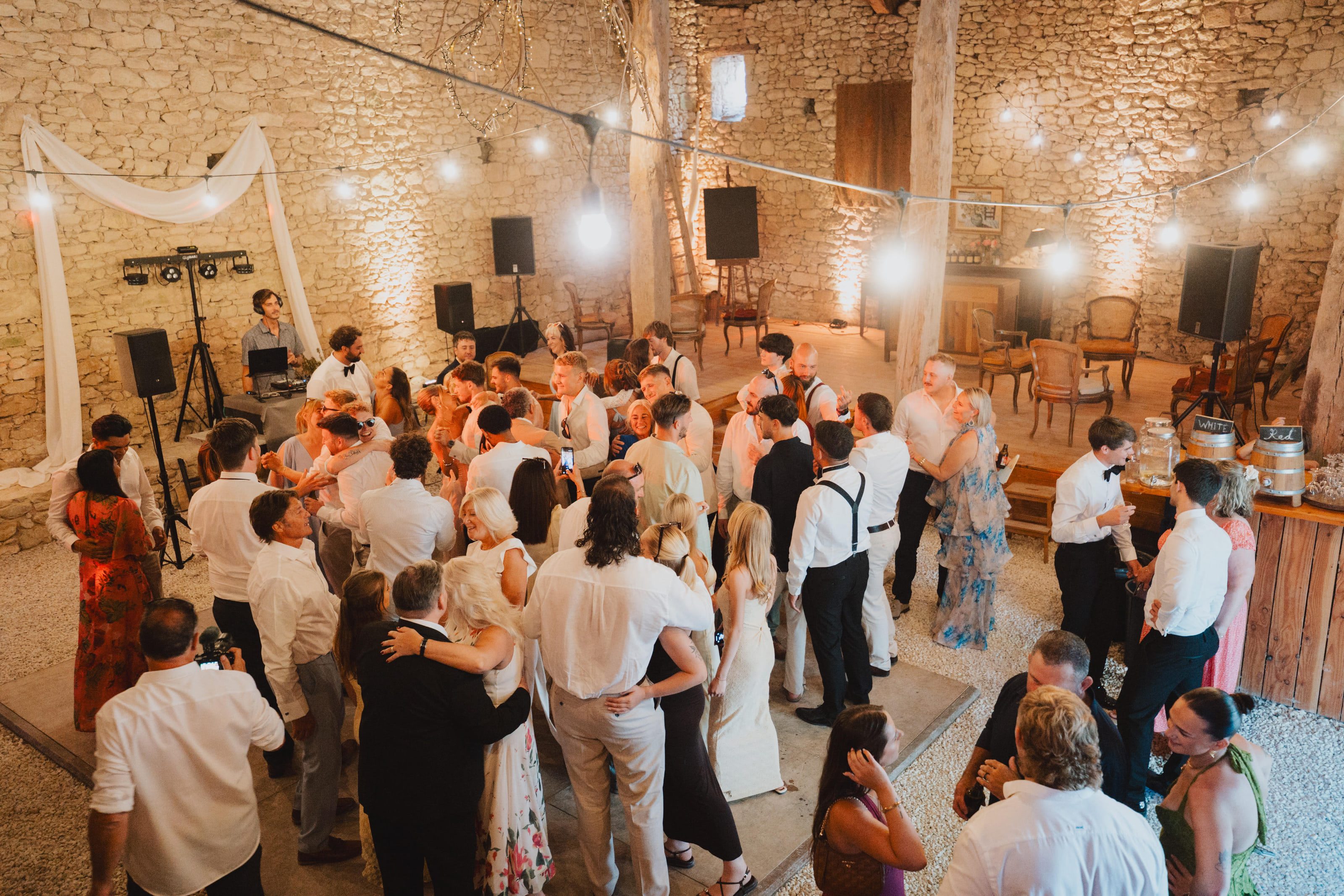 A full dance floor in a stone barn, festoon lights overhead and the DJ booth at the far end.