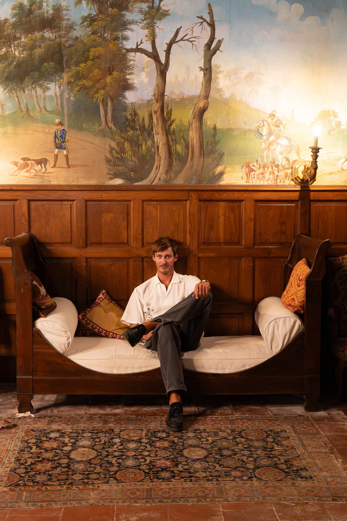 Denver seated on a château daybed beneath a painted landscape mural, by candlelight.